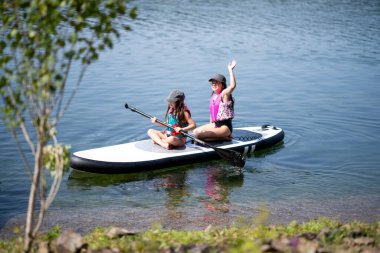 Sahile yakın SUP panosunda iki kız, kameraya el sallayan bir el, fotokopi alanı ile eğlenceli yaz çocukluk macerası.