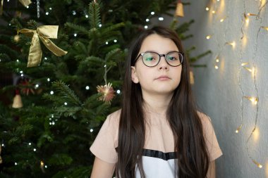 Gözlüklü ve tişörtlü üzgün genç kız, süslü Noel ağacının yanında, duygusal çocukluk konsepti ve sakin şenlik havası
