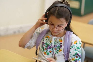 Gözlüklü, nakışlı tişörtlü, kulaklıkla dinleyen ve elinde akıllı telefon, modern eğitim teknolojisi konsepti olan bir kız öğrenci.