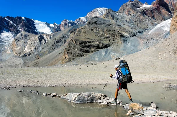 Un homme avec un sac à dos reposant sur un bâton de trekking déplace une rivière — Photo