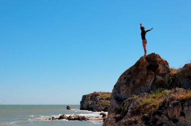 Deniz mavi gökyüzü karşı düşünürken ayakta ve doğal kıyı kaya, yüksek üzerinde rahatlatıcı kadın.