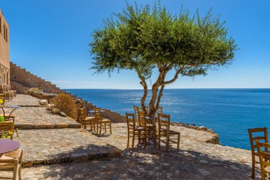 Traditional cafe exterior in the fortified medieval  castle of Monemvasia. Iron tables and wooden chairsand an olive tree  with the view of the  aegean sea in the background. clipart