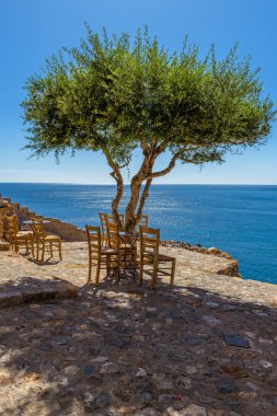 Monemvasia 'nın güçlendirilmiş ortaçağ şatosunda geleneksel bir kafe. Demir masalar, ahşap sandalyeler ve arka planda Ege Denizi manzaralı bir zeytin ağacı..