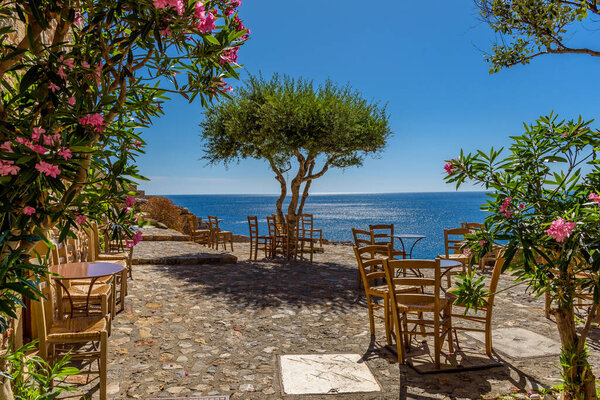 Traditional cafe exterior in the fortified medieval  castle of Monemvasia. Iron tables wooden chairs and an olive tree  with the view of the  aegean sea in the background.