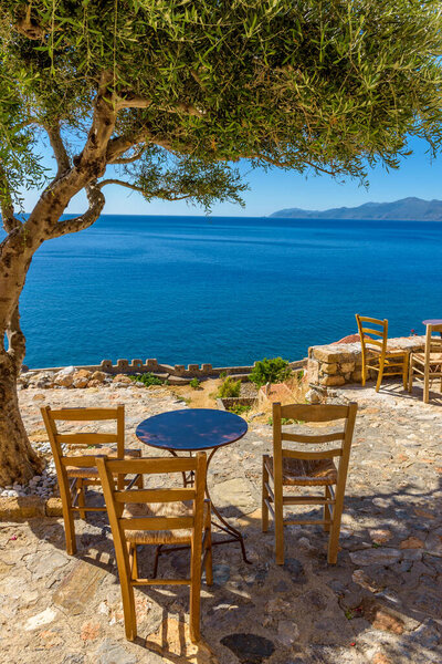 Traditional cafe exterior in the fortified medieval  castle of Monemvasia. Iron tables and wooden chairsand an olive tree  with the view of the  aegean sea in the background.