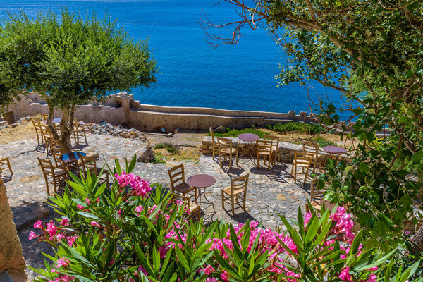 Traditional cafe exterior in the fortified medieval  castle of Monemvasia. Iron tables and wooden chairs and an olive tree  with the view of the  aegean sea in the background.