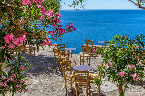Traditional cafe exterior in the fortified medieval  castle of Monemvasia. Iron tables and wooden chairs and an olive tree  with the view of the  aegean sea in the background.