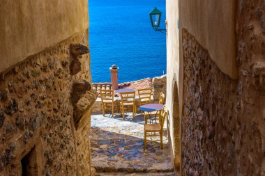 Monemvasia 'nın güçlendirilmiş ortaçağ şatosunda geleneksel bir kafe. Demir masalar ve arka planda Ege Denizi manzaralı ahşap sandalyeler.