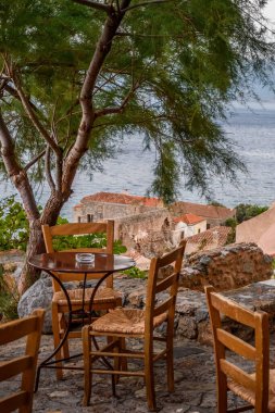 Monemvasia 'nın güçlendirilmiş ortaçağ şatosunda geleneksel bir kafe. Demir masalar ahşap sandalyeler ve arka planda Ege Denizi manzaralı bir zeytin ağacı..