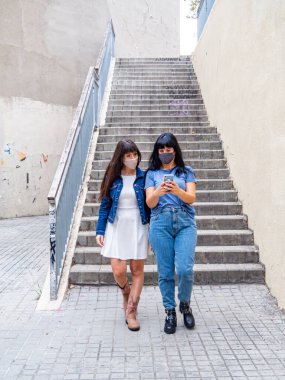 Kafkas kökenli iki genç kadın caddede yüzlerinde maskelerle yürüyorlar ve cep telefonuna bakıyorlar. Arkadaşlık, milenyum ve koronavirüs konsepti. Arka plandaki merdivenler..