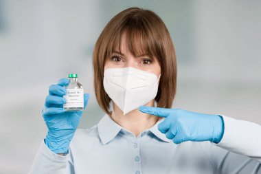 Yüz maskesi ve eldivenlerle korunan kadın doktor veya hemşire elinde bir koronavirüs / covid aşı şişesini işaret ediyor.