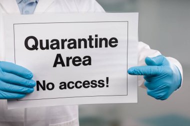 Doktorun elinde tıbbi eldiven var. Klinik odasının önünde karantina işareti var.