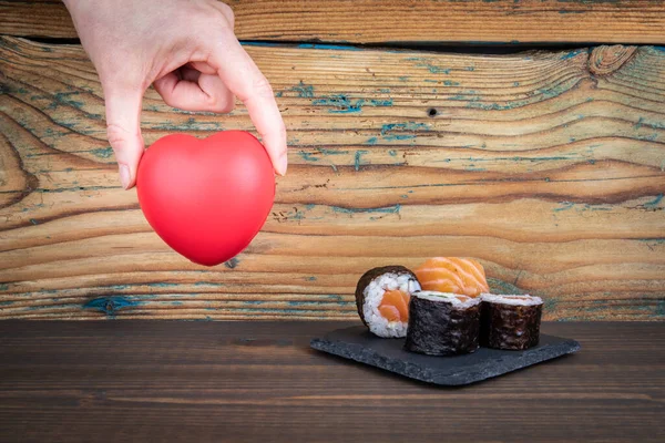 Červené gumové srdce v ženské ruce. Jídlo, vitamíny a kultura. Sushi rolka s lososem — Stock fotografie