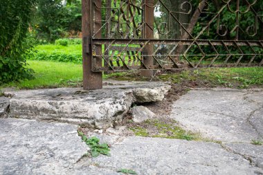 Paslı bir direğe bağlanmış yıpranmış metal bir kapı bir bahçe girişindeki kırık ve harap beton zeminde duruyor..