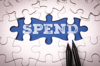 Beyaz bulmaca parçalarının altında mavi arka plan üzerine yazılan kelime harcamaları para yönetimi ve yatırım planlama kavramını sembolize ediyor.