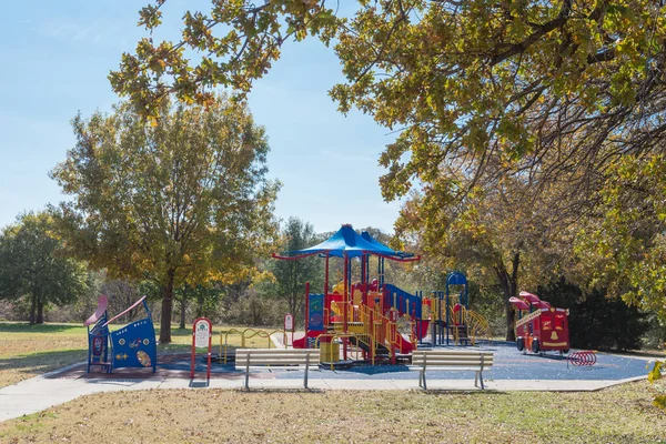 Two empty metal benches looking at vibrant playground surrounded with colorful autumn leaves in Flower Mound, Texas, America. Public playground with shade sail and rubber mat flooring