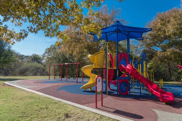 Close-up red slide tube of vibrant playground with colorful fall foliage in Flower Mound, Texas, America. Public playground with shade sail and rubber mat flooring near nature park