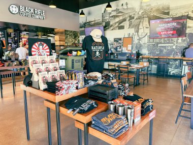 NORTH RICHLAND HILLS, TX, US-OCT 4, 25: Branded merchandise including mugs, hats, and apparel at Black Rifle Coffee Company with customer placing drink order at counter, surrounded by warm lighting