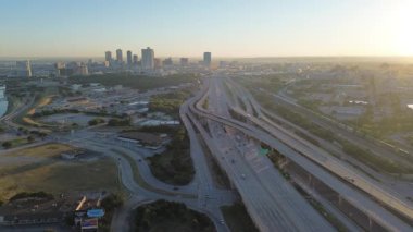 Fort Worth four-level stack interchange near downtown, with I-30 and I-35W converging beside the Trinity River. Sunrise light reveals layered ramps, skyline silhouettes, busy commuter flow, Texas
