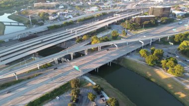 Busy highway traffic and early morning runners, walkers trace Trinity Trails beside river, elevated channel toward Fort Worth hospital district. Green buffers soften the city infrastructure edge