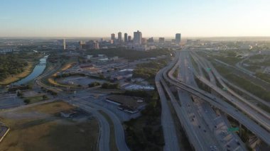 Curved Trinity River and stacked highway system lead into downtown Fort Worth, framed by light industrial buildings in foreground. Early morning light adds warmth to city layered commute corridor