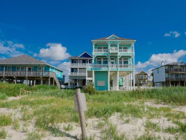 Plaj uyarı levhası, çimenli sahil şeridi üzerindeki tahta tehlike işareti iki katlı sahil evi, beyaz süsleme, veranda mobilyası, açık kiriş. Balkonlar rahatlamaya davet eder, Galveston, TX. ABD