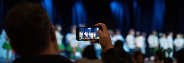 Panorama View 'daki adamın elinde akıllı telefon kayıtları var. Çocuklar sahnede Noel şarkıları söylüyorlar. Bulanık sahneyle tamamen zıtlar. Toplumsal deneyimlerin dijital korunumu, Teksas. ABD