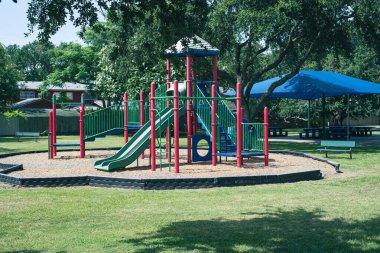 Yeşil kaydırak, kırmızı direkler ve yeşil raylarla çerçevelenmiş mavi çatılı platformlardan iniyor. Ağaç parçaları, siyah kenarlar ve piknik tezgahları Galveston, Teksas 'taki yemyeşil ağaç örtüsünün altında. ABD