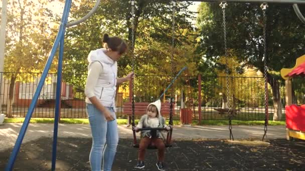 Щаслива мама кидає маленьку дитину на гойдалці в парку. Мати з донькою ходить у вихідні. Сімейна прогулянка в парку. Хлопчик і юний батько. Спільна робота — стокове відео