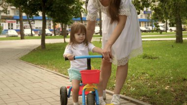 Sağlıklı çocuk bisiklet sürmeyi öğrenir. Ebeveynler ve küçük kızları parkta yürürler. Mutlu anne küçük kızına bisiklet sürmeyi öğretiyor. Anne bebeğiyle dışarıda oynuyor. Mutlu aile ve çocukluk kavramı