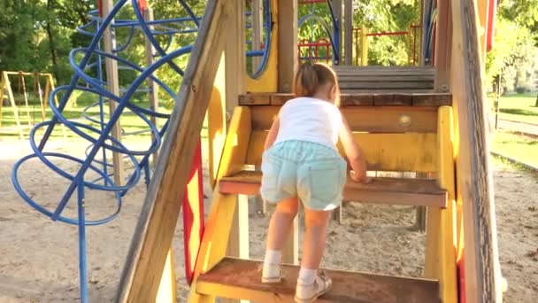 Gelukkige kindertijd concept. kind klimt de trap op naar kinderglijbaan. Moeder en baby spelen op de speelplaats. Gelukkige kindertijd en familie concept. klein kind lacht en geniet van speeltuin in het park — Stockvideo