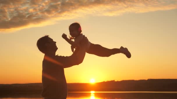 Papà getta la sua figlioletta felice in aria sulla spiaggia, divertendosi al tramonto. padre gioca con il bambino con l'acqua. felice concetto di famiglia. silhouette di un padre e un bambino sano che sorvola il sole — Video Stock