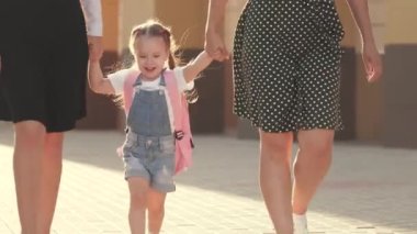 Sağlıklı küçük bir kız, annesi ve kız kardeşiyle oynar, ellerini tutar ve zıplar. Aile ve çocukluk kavramı. Neşeli çocuk atlaması. Mutlu aile, anne ve çocuklar okula gidiyor.