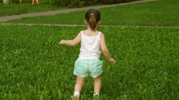 Boldog kislány fut a zöld füvön egy nyilvános parkban egy nyári napon. A gyermek a friss levegőn sétál és játszik. Sétáló kölyök. Gyönyörű mező a hőségben. — Stock videók
