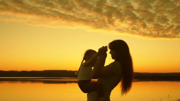 Mamma och dotter leker vid solnedgången. Mor håller ett lyckligt barn i sina armar mot bakgrunden av himlen. Lycklig familj koncept. Grabben och kvinnan på en kvällspromenad. Silhuett — Stockvideo