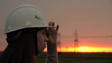 Beyaz kasklı güzel bir mühendis kız gün batımında elektrik hattına bakıyor. Gökyüzüne karşı enerjik bir kadın. Dağıtım ve elektrik tedariği. Temiz enerji. Yakın plan.