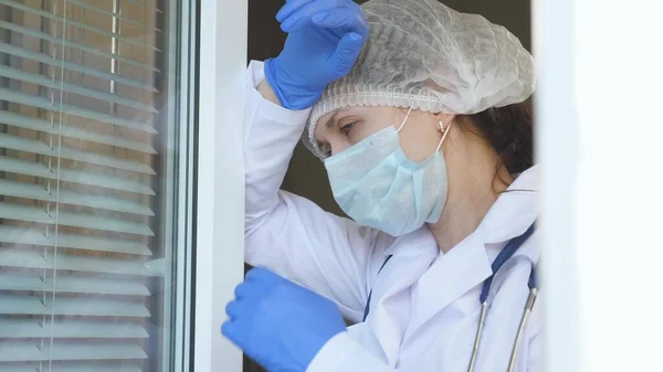 Une femme médecin fatiguée en combinaison de protection s'approche de la fenêtre, enlève son masque médical et son chapeau, ferme les yeux de la fatigue, respire de l'air frais. Dur journée de travail du médecin à l'hôpital . — Photo