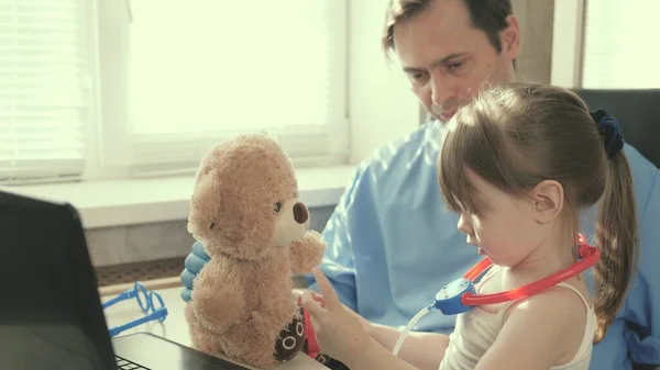 Zorgvuldige professionele mannelijke kinderarts spelen met een klein kind in het kantoor. klein meisje speelt met teddybeer speelgoed luistert naar hem met een stethoscoop. peuter patiënt zit op schoot van kinderarts. — Stockfoto