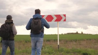Sırt çantalı gezginler kavşaktaki yol işaretine gelip rotalarını seçiyorlar. Turistlerin takım çalışması. Yürüyüş ve macera konsepti. Erkekler kaybolur, yolculuk için doğru yönü ararlar..