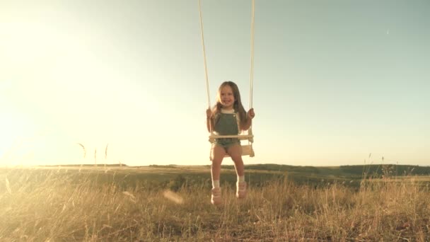 Niño feliz vuela en un columpio de cuerda en el parque. niña alegre ríe, se regocija y agita los brazos en vuelo. Mamá sacude a su hija sana en un columpio bajo el árbol bajo el sol. infancia y familia. — Vídeos de Stock