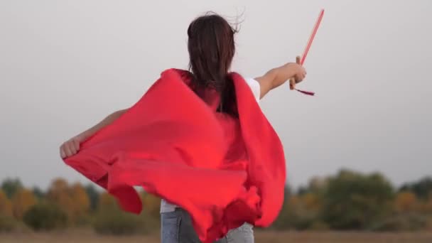 Leka riddare. gratis flicka i röd mantel körs med svärd i handen över fältet spelar medeltida riddare. En ung flicka som spelar superhjältar. Barn slåss med leksakssvärd. lycklig barndom — Stockvideo