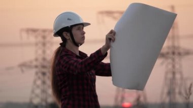 Bir mühendisin silueti yüksek voltajlı bir elektrik santrali inşa projesine bakıyor. Mühendis çizimi okuyor. Mühendis gün batımında elektrik direklerinin yanındaki alanda çalışıyor..