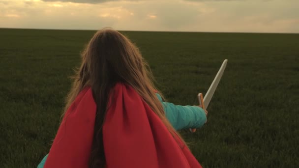 Flicka i en röd mantel löper med svärd i handen över fältet spelar medeltida riddare. En ung flicka som spelar superhjältar. Leka riddare. Barn slåss med ett leksakssvärd. Glad barndom. — Stockvideo
