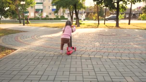 Gesundes Kind fährt Roller in der Stadt auf der Straße. Glückliche Kindheit. Kleines Mädchen lernt Rollerfahren. glückliches kleines Mädchen spielt im Park. — Stockvideo