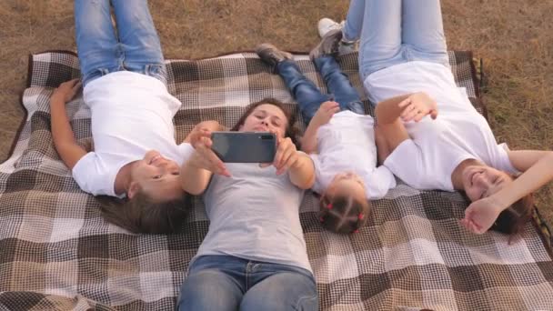 Mamma e bambini felici vengono fotografati sullo smartphone nel parco. Madre e figlie si fanno un selfie mentre viaggiano, sdraiate sul copriletto. famiglia felice e smartphone. divertenti bambini e genitori in vacanza — Video Stock