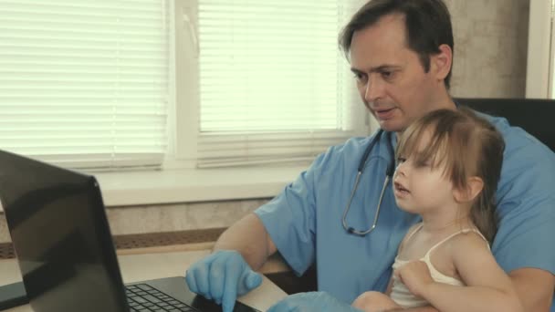 Koncepcja badania pediatrycznego w leczeniu dzieci. Opiekuńczy profesjonalny lekarz pediatra, bawiący się uroczym dzieckiem w przedszkolu i pracujący na laptopie. dziewczyna pacjent siedzi na kolanach pediatra — Wideo stockowe