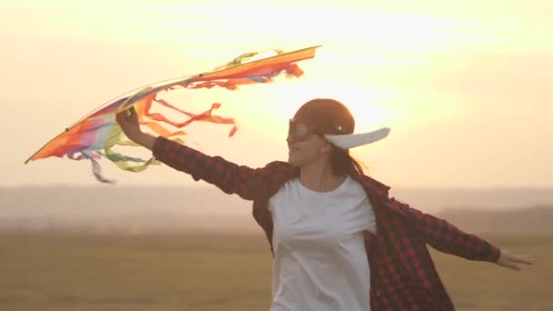 Happy girl runs with a multi-colored kite in her hands to field in rays of sun. teenager wants to become pilot. child dreams of freedom, flight and a happy family. teenager plays by airplane outdoors. — Stock Video