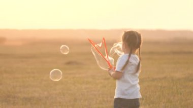 Sahada sabun köpükleri, çocukların doğum günü. Sağlıklı çocuk parkta sabun köpükleriyle oynuyor. Bebek hava kabarcıklarında seviniyor. Kızı gün batımında parkta oynuyor. Mutlu aile ve çocukluk kavramı.