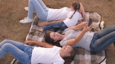 Anne ve mutlu çocuklar parkta akıllı telefondan fotoğraf çekiliyor. Anne ve kızları seyahat ederken yatak örtüsünde uzanırken selfie çekiyorlar. Mutlu aile ve akıllı telefon. Komik çocuklar ve tatilde ebeveynler