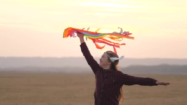 Happy child in a pilots helmet runs with a multi-colored kite in his hands at sunset. girl wants to become pilot. child dreams of freedom, flight and travel. teenager playing on a plane outdoors. — Stock Video